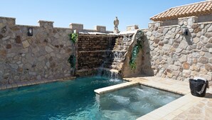 Uma piscina com paredes de pedra, com uma cascata, um banco de pedra e uma parede de pedra com uma pequena janela.