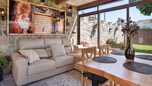 Uma sala de estar acolhedora com parede de pedra, sofá bege, mesa de jantar de madeira e uma janela grande com vista para uma parede de pedra e vegetação.