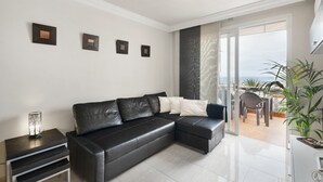 A modern living room with a black leather sofa, a small side table, and a large window offering a view of the outdoors. - Vacation Apartment 'The Sunset' with Shared Pool, Wi-Fi and Air Conditioning (Costa Adeje)