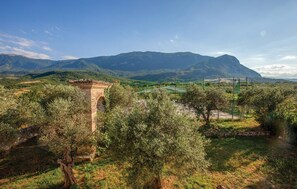 Une arche en pierre se dresse dans un paysage rural, avec des oliviers et une montagne en arrière-plan.