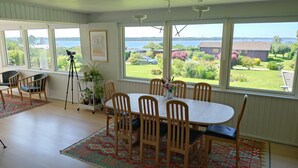 A dining area with a table and chairs, a tripod, and a view of a lake and greenery. - Cozy summerhouse with spectacular views! (Ølsted)