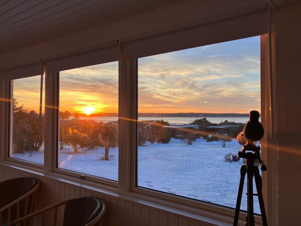 Ein Zimmer mit einem Teleskop auf einem Stativ, großen Fenstern und Blick auf eine verschneite Landschaft bei Sonnenuntergang.