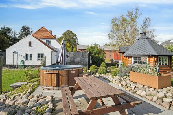 A wooden deck with a hot tub, picnic table, and a stone pathway leading to a house with a red roof.