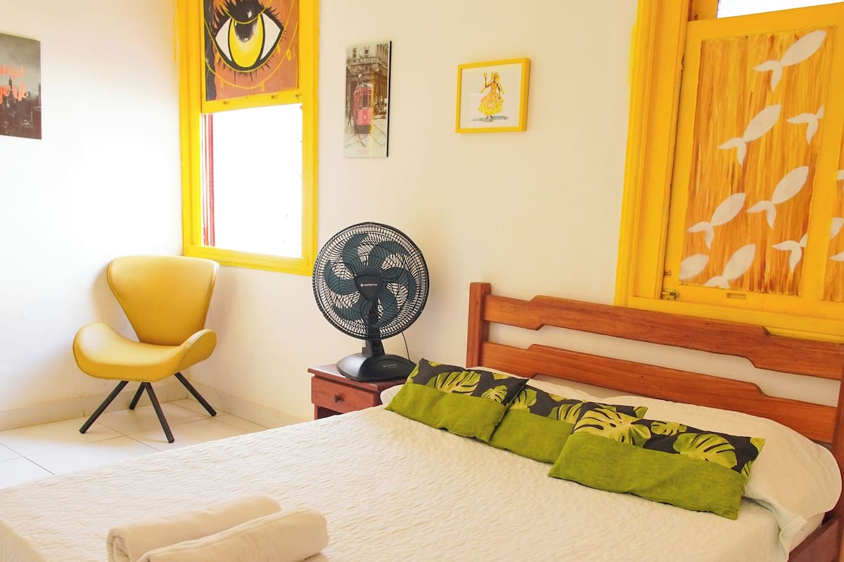 A neatly made bed with a white bedspread and green pillows, a yellow chair, a bedside table with a fan, and a window with yellow blinds.