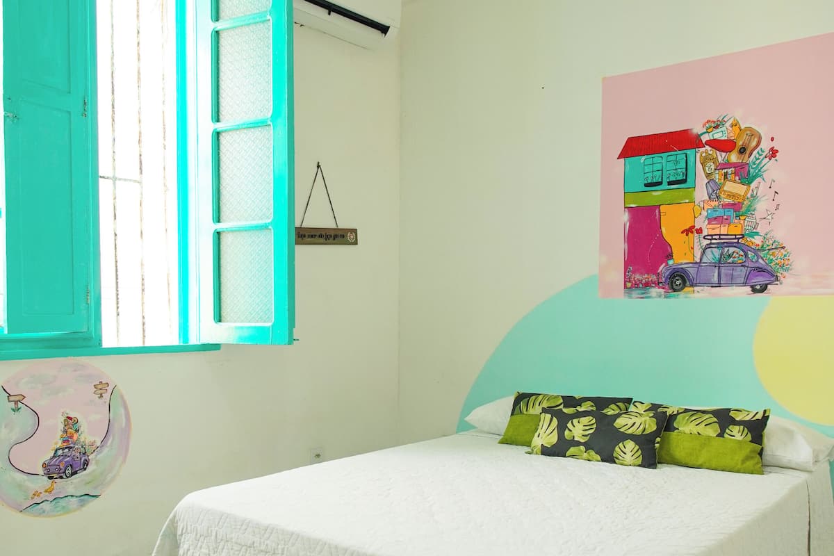 A bedroom with a bed, a window with blue shutters, a wall with a colorful painting, and a wall-mounted air conditioner.