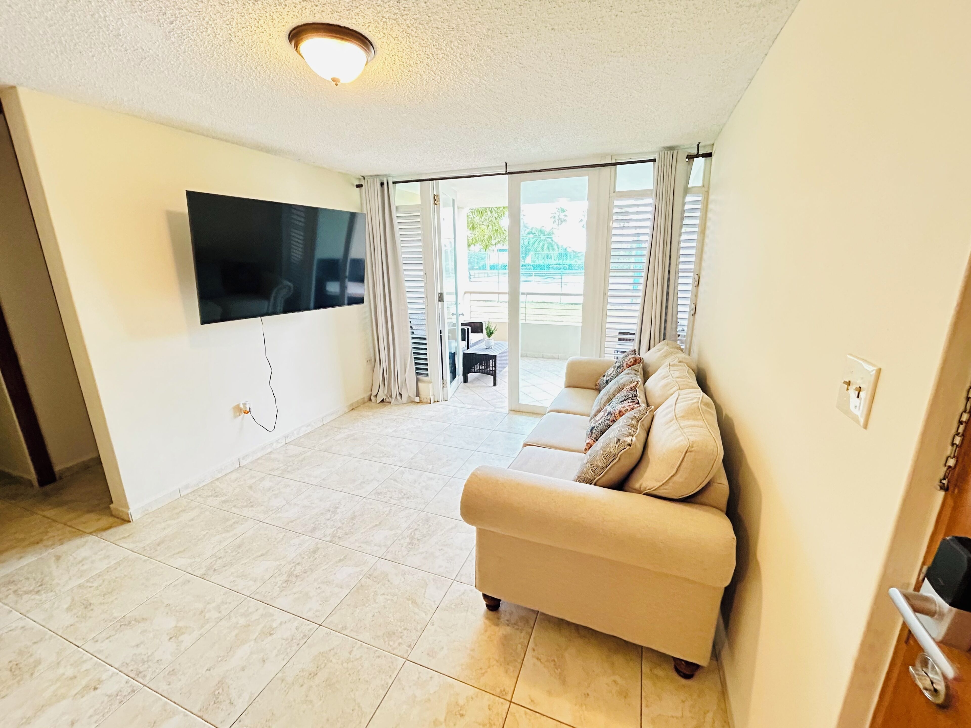 A spacious living room with a flat-screen TV mounted on the wall, a beige sofa with patterned cushions, and a sliding glass door leading to an outdoor area.