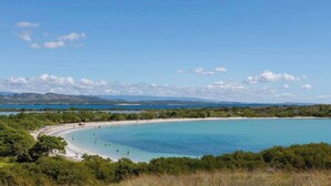 Een kuststrand met helder turquoise water, een zandstrand en een achtergrond van groene heuvels en bergen.