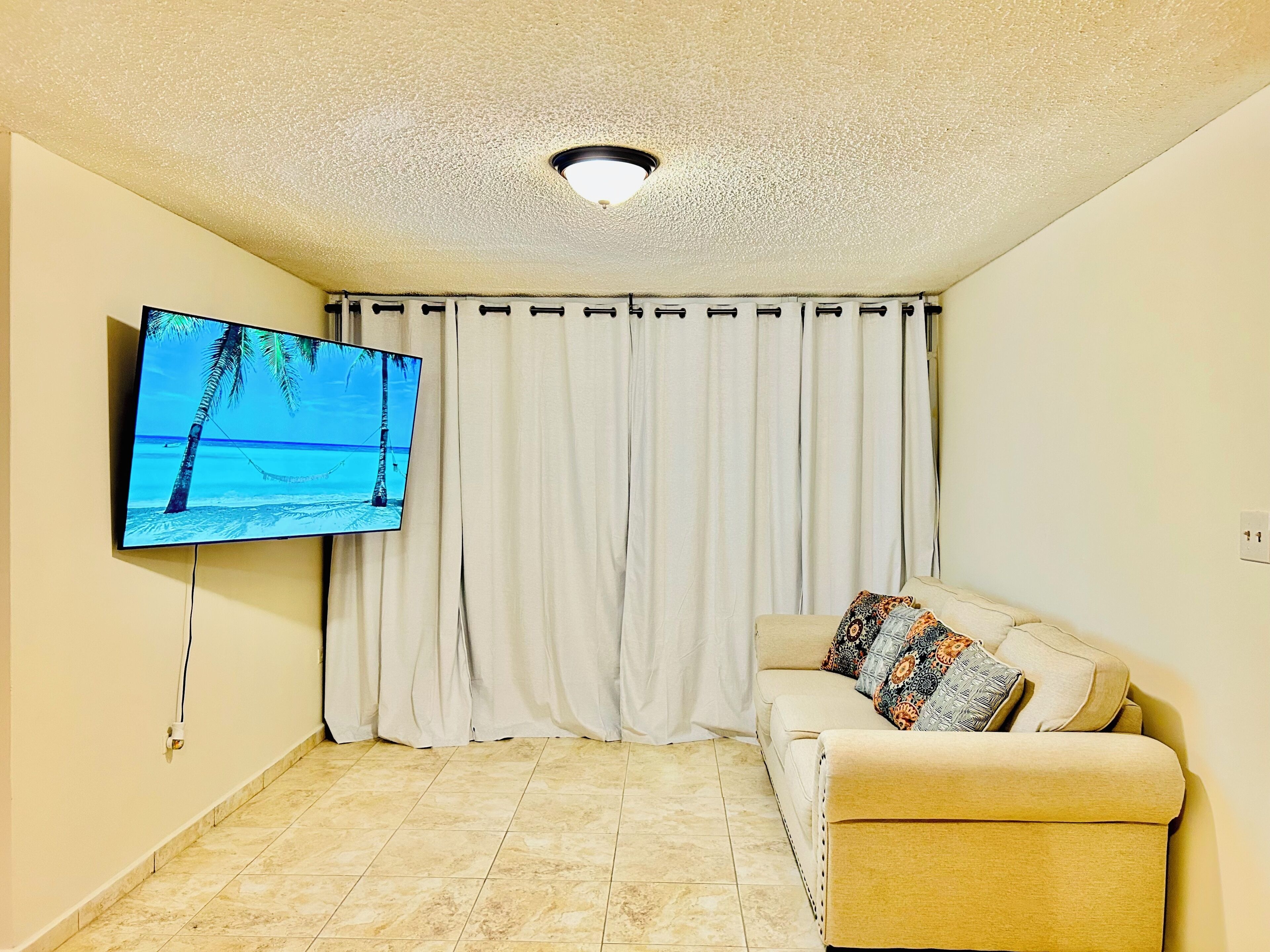Una sala de estar con una televisión de pantalla plana en la pared, un sofá color beige con cojines estampados, una cortina blanca y un piso de baldosas.