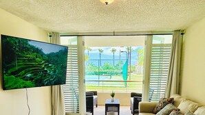 Een ruime woonkamer met een grote flatscreen televisie aan de muur, een beige bank met gekleurde kussens, een kleine tafel met een plant in pot en uitzicht op een tennisbaan door grote ramen.