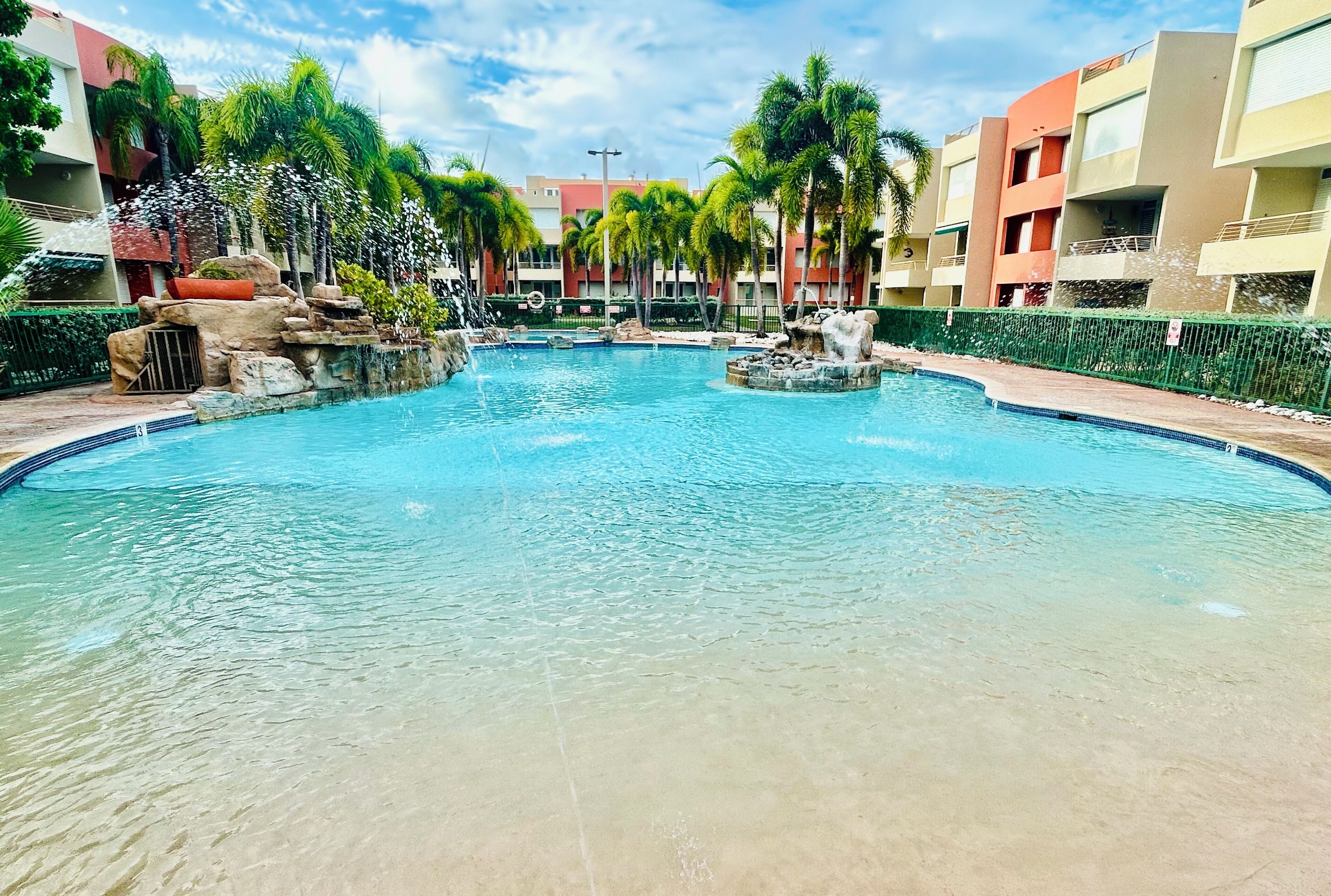 A swimming pool with clear water, surrounded by palm trees and a building with balconies.
