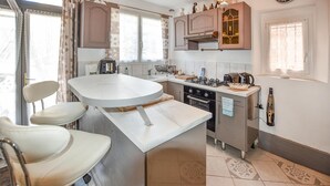 A compact kitchen with a central island, a built-in oven, and a gas stove. There are two bar stools and a window with patterned curtains. - 1 bedroom cozy home in Moustiers-Sainte-Marie (Moustiers-Sainte-Marie)