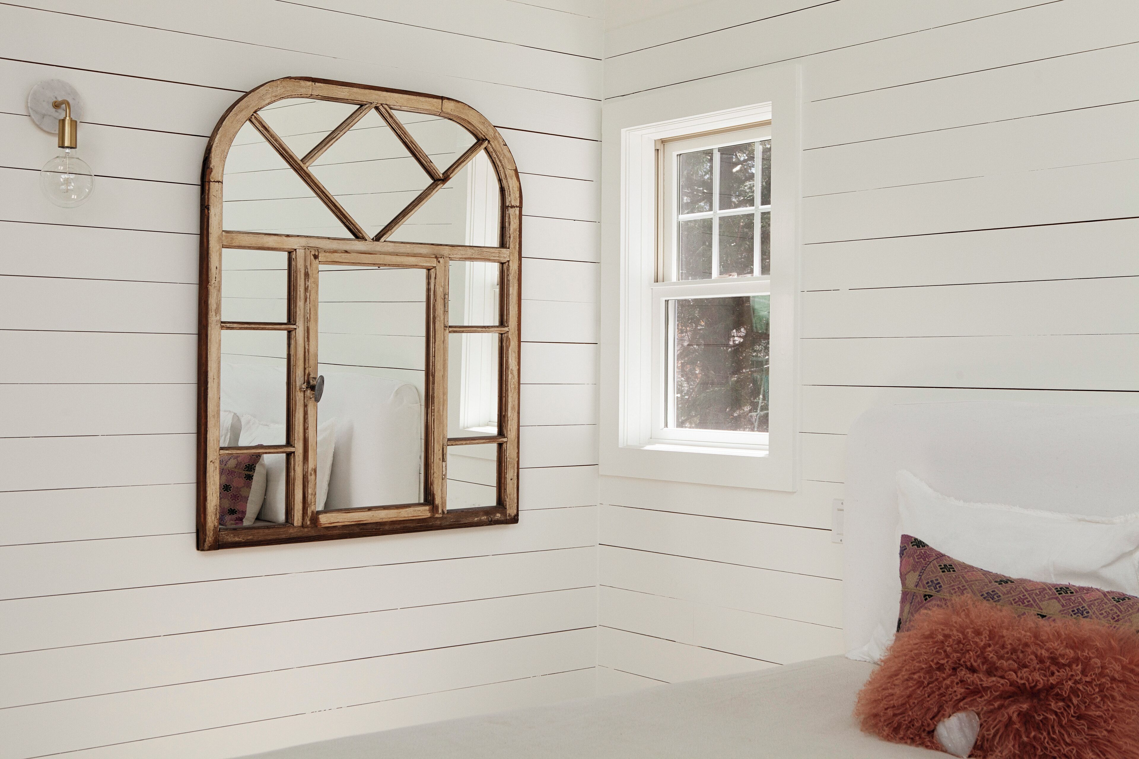 A room with white wooden paneling, a large arched mirror, a window, and a fluffy pillow.