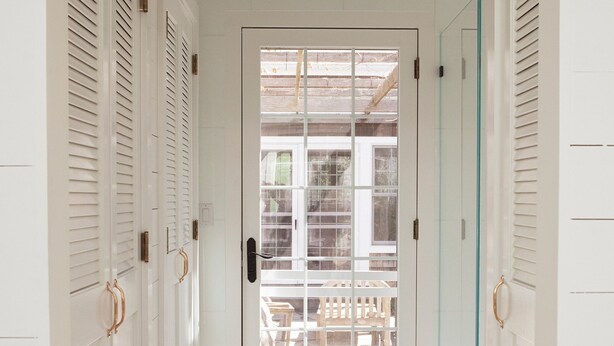 Un couloir avec des portes blanches, une porte vitrée donnant sur l’extérieur et un plancher en bois.