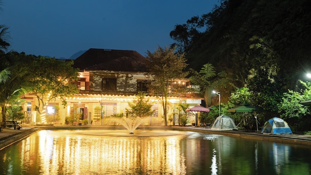 A nighttime view of a hotel with a fountain in the courtyard, surrounded by trees and a pool. - Wind's House Ha Giang (Vi Xuyen)
