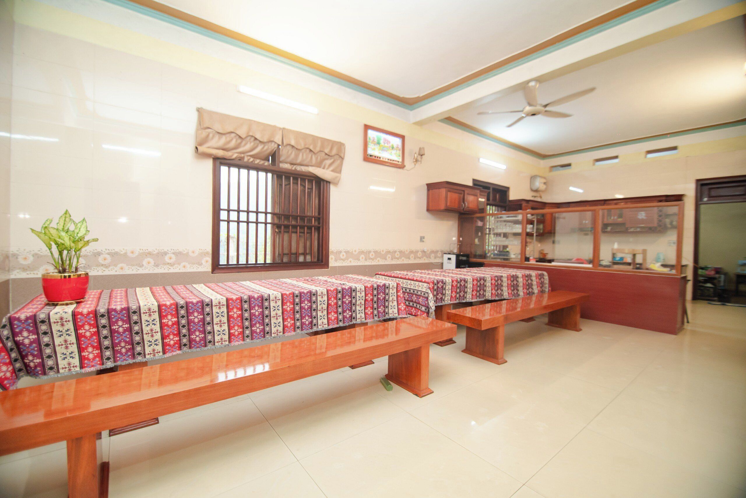 A room with wooden benches covered with patterned tablecloths, a potted plant, a window with bars, a ceiling fan, and a display cabinet.