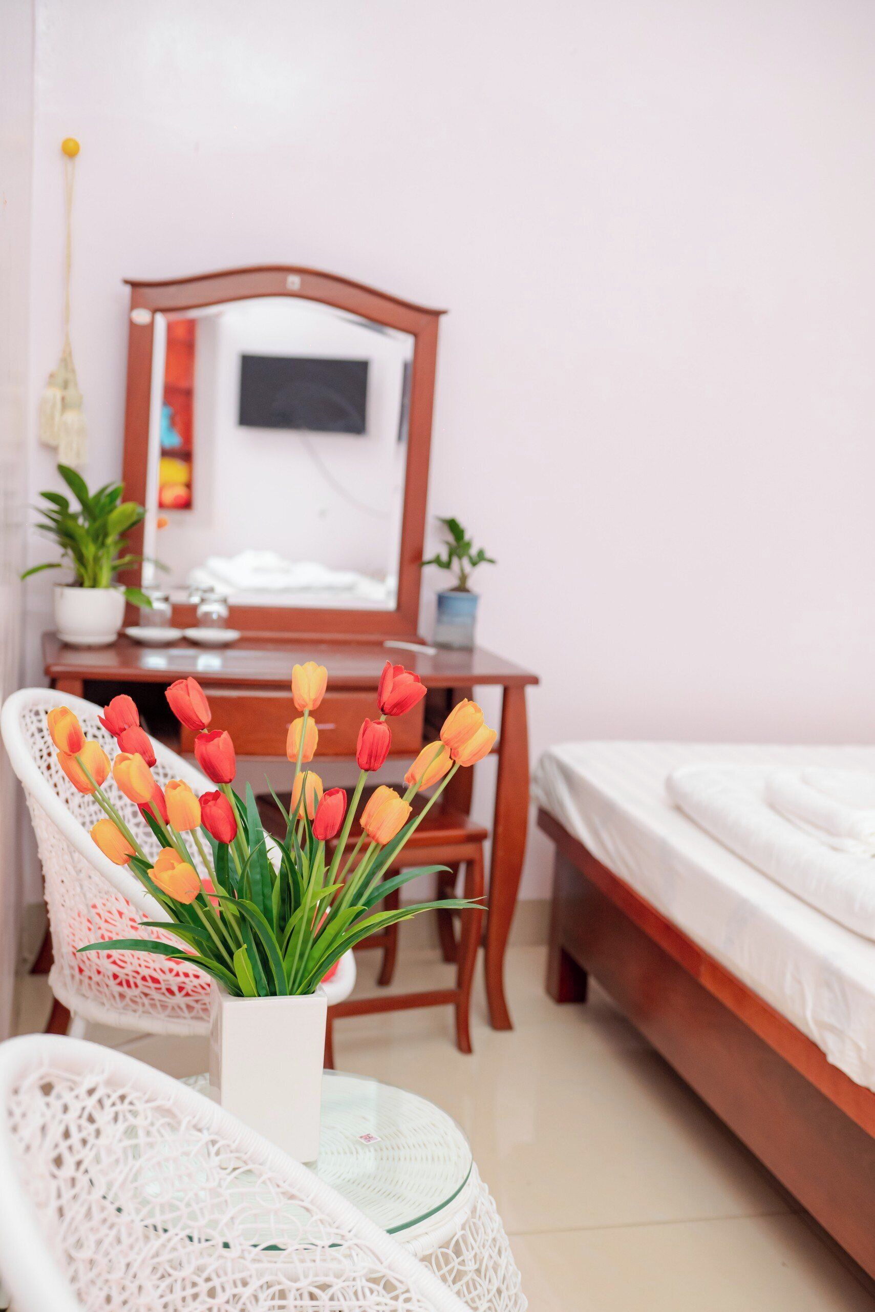 Een netjes ingerichte slaapkamer met een houten bed, een nachtkastje, een stoel en een bureau met een spiegel. Op het bureau staat een vaas met tulpen.