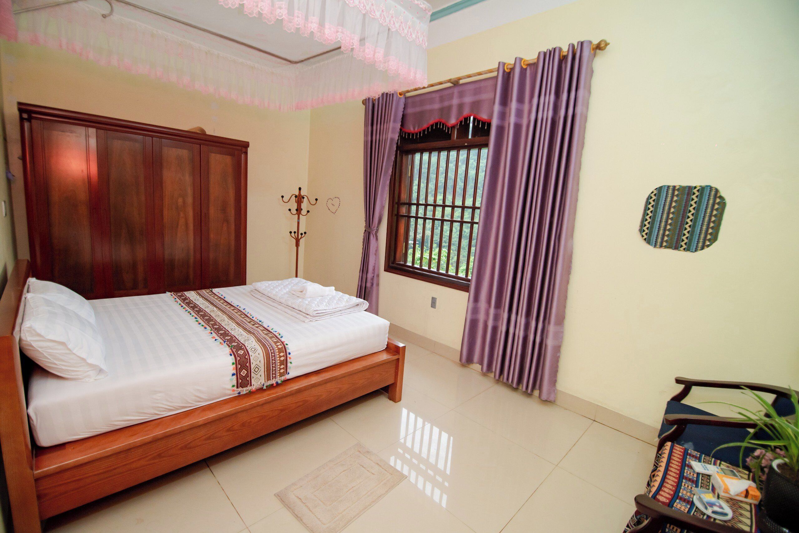 A neatly made bed with a wooden headboard, a window with purple curtains, a small table with a plant, and a decorative wall piece.