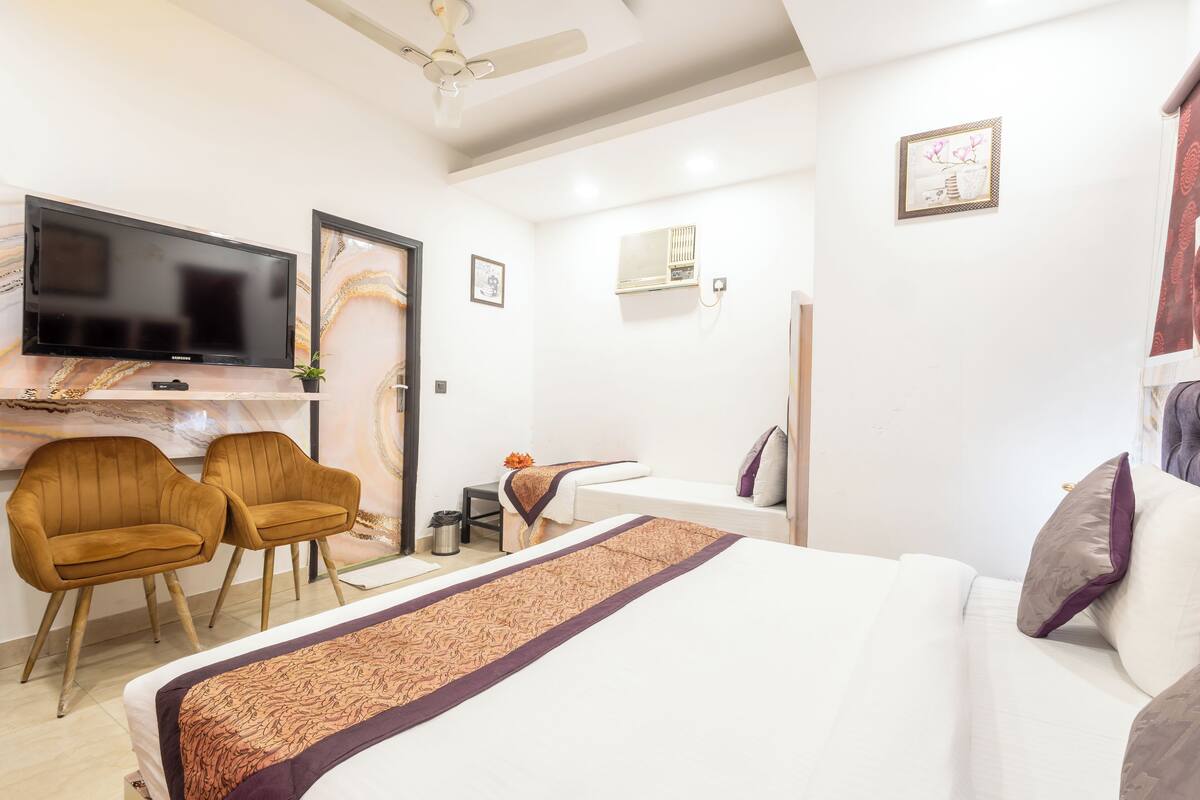 A hotel room with a bed, two armchairs, a desk with a flat-screen TV, a ceiling fan, and a small table with a vase.