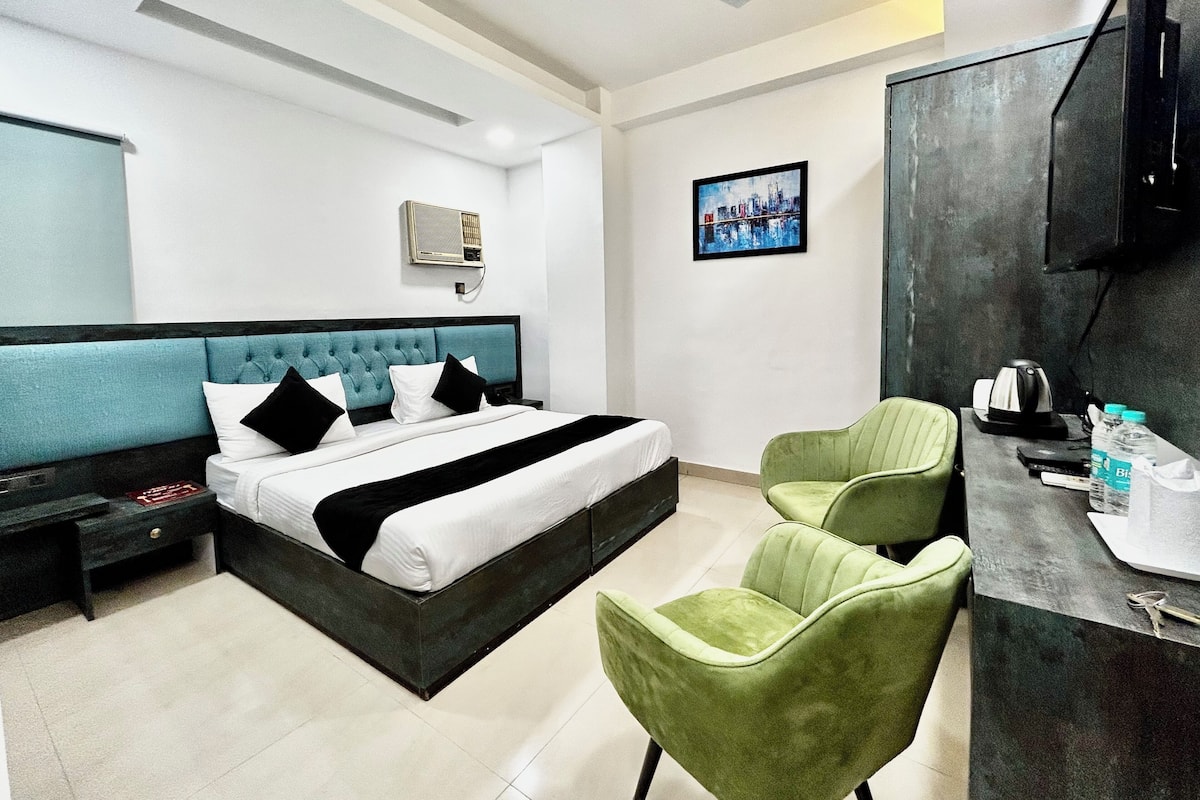 A modern hotel room with a bed, a desk, two green chairs, a ceiling fan, a picture on the wall, and a water bottle on the desk.