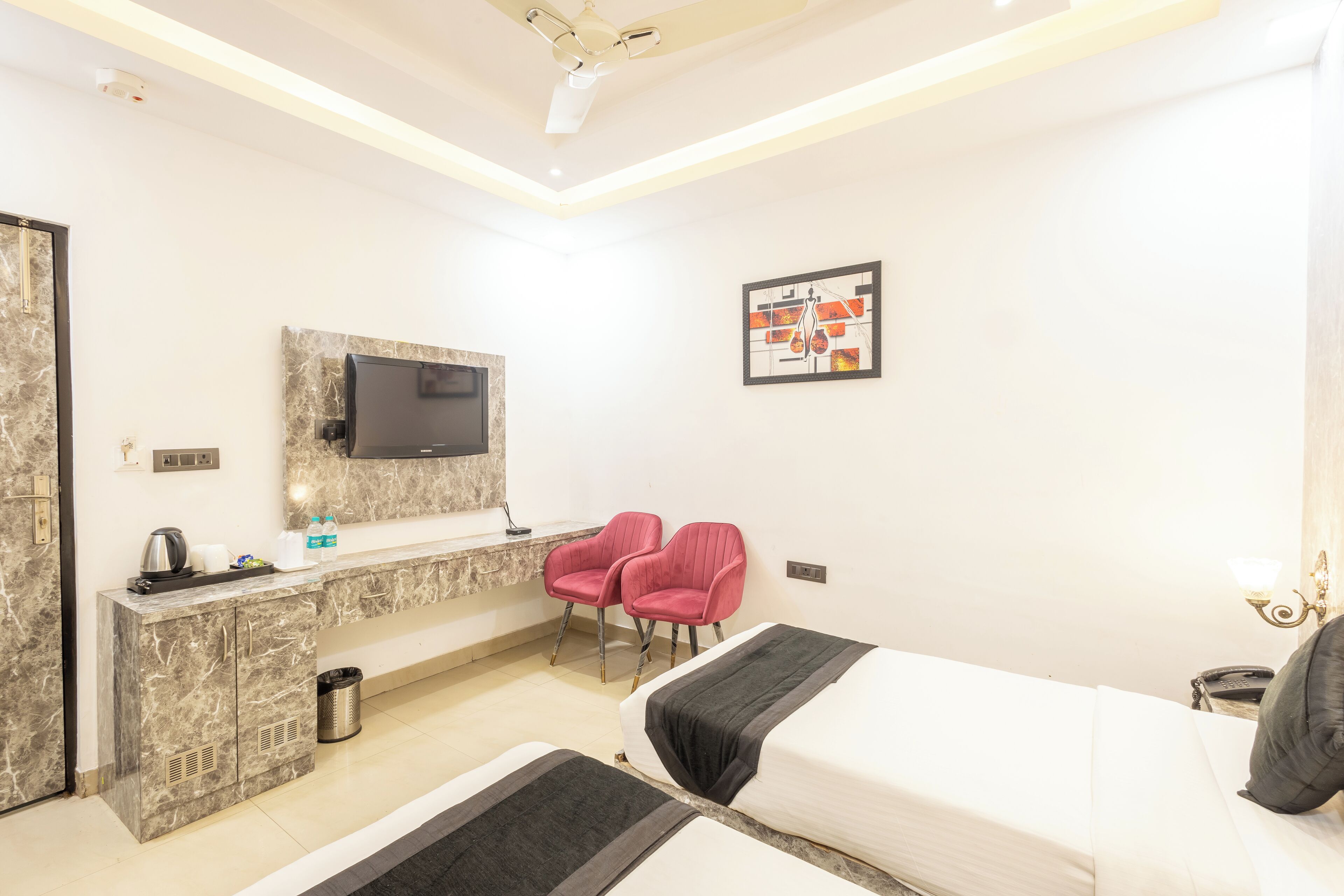 A hotel room with a bed, a desk with a TV, two red chairs, and a small trash bin.