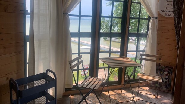 Una habitación acogedora con un amplio ventanal, una mesa de madera y dos sillas. Se observa un cartel de bienvenida en la pared.