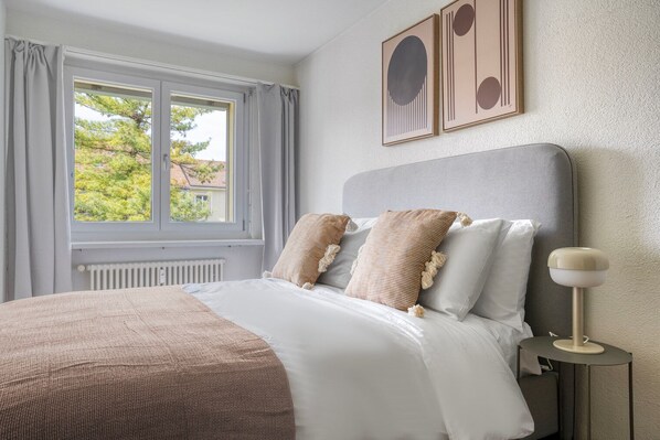 A neatly made bed with a grey headboard, a bedside table with a lamp, and a window with curtains.