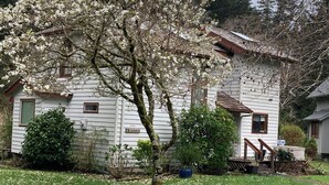A white house with a green door, surrounded by a garden with blooming trees. - Riverfront Beach House Rental (Yachats)