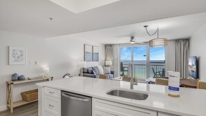 A modern kitchen with white cabinetry, a built-in dishwasher, and a large island with a sink. There is a dining area with a table and chairs, and a view of the ocean through large windows.