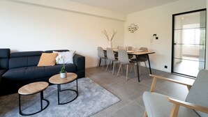 A modern living room with a dark blue sofa, two round tables, and a dining area with a glass door leading outside. - 't Zout with balcony by Interhome (Bredene)