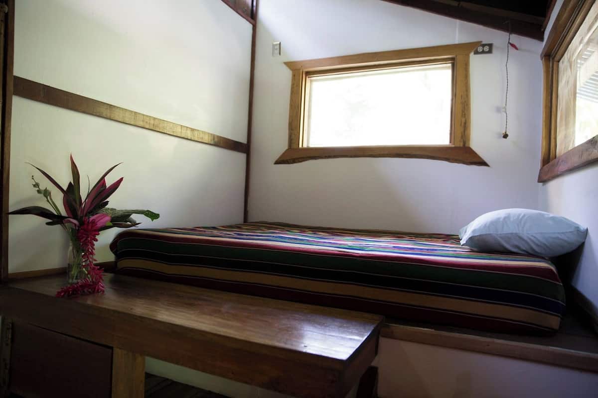 A small, cozy room with a single bed, a wooden bench, and a window providing natural light.
