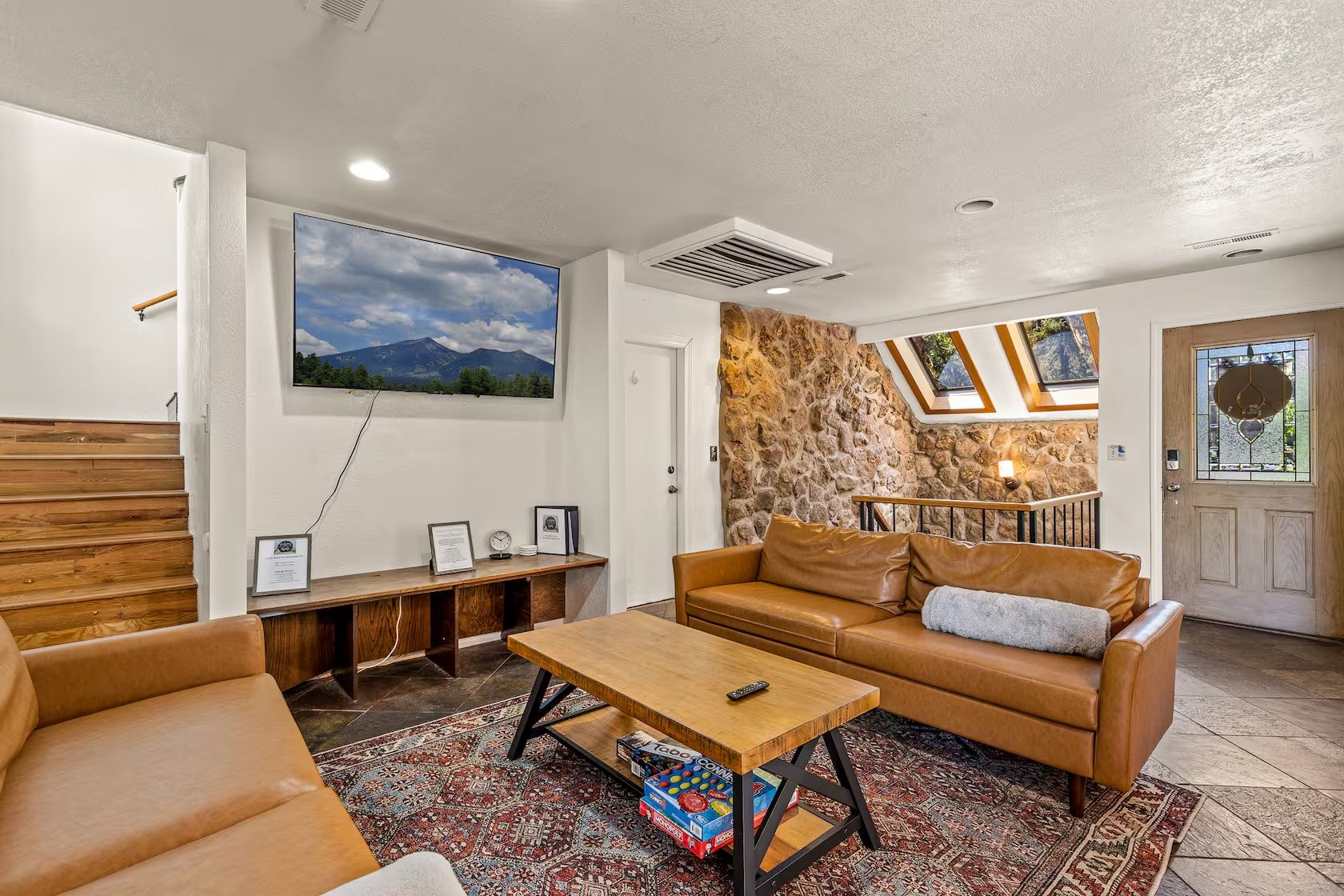 Uma sala de estar com um banco de madeira, um sofá de couro, uma mesa de centro e uma TV fixada na parede.