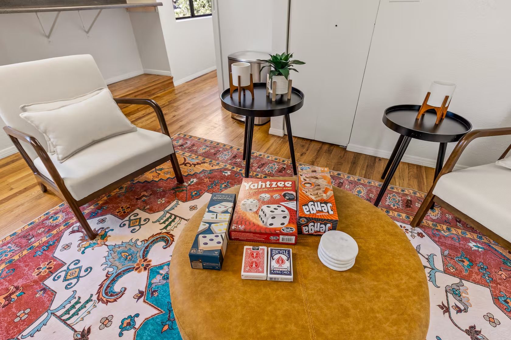 Uma sala de estar com um tapete estampado, duas cadeiras brancas, uma mesa de centro redonda e uma mesinha lateral com uma planta.
