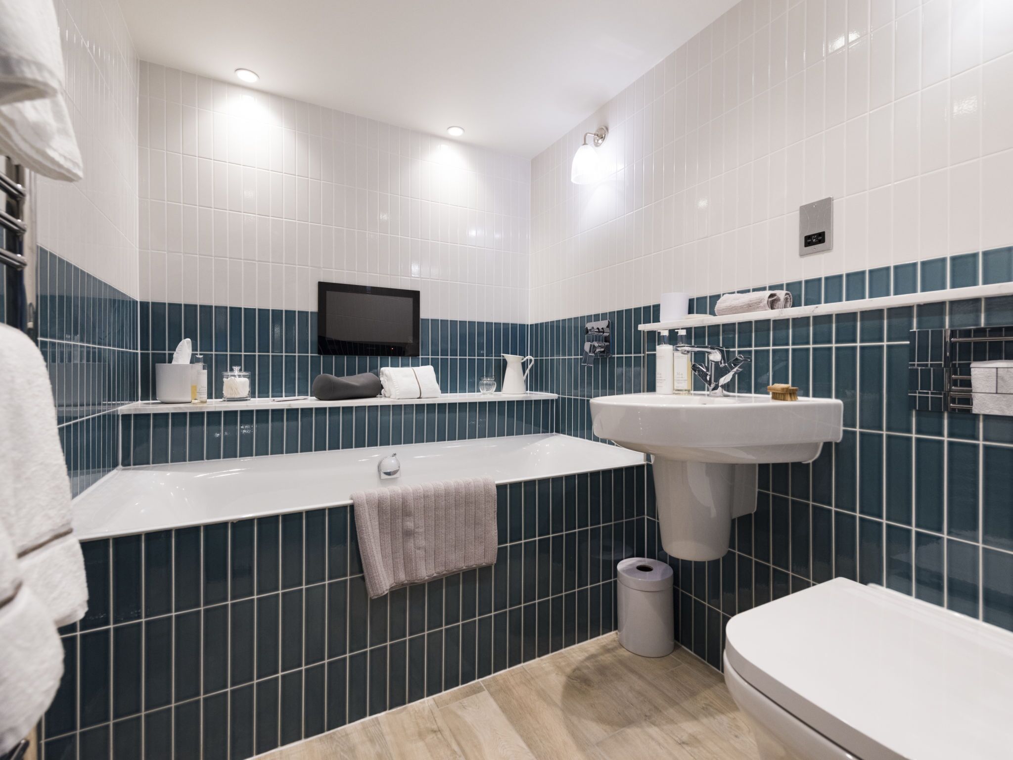 A modern bathroom with a bathtub, sink, and toilet. There is a TV mounted on the wall and a towel rack.