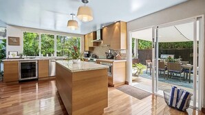 Une cuisine moderne dotée d’un îlot central, de meubles en bois et d’une grande porte-fenêtre coulissante donnant sur un espace repas extérieur.
