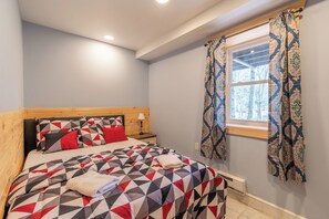 Une chambre à coucher avec une couvre-lit à motifs géométriques, une tête de lit en bois, une table de chevet avec une lampe et une fenêtre ornée de rideaux à motifs.