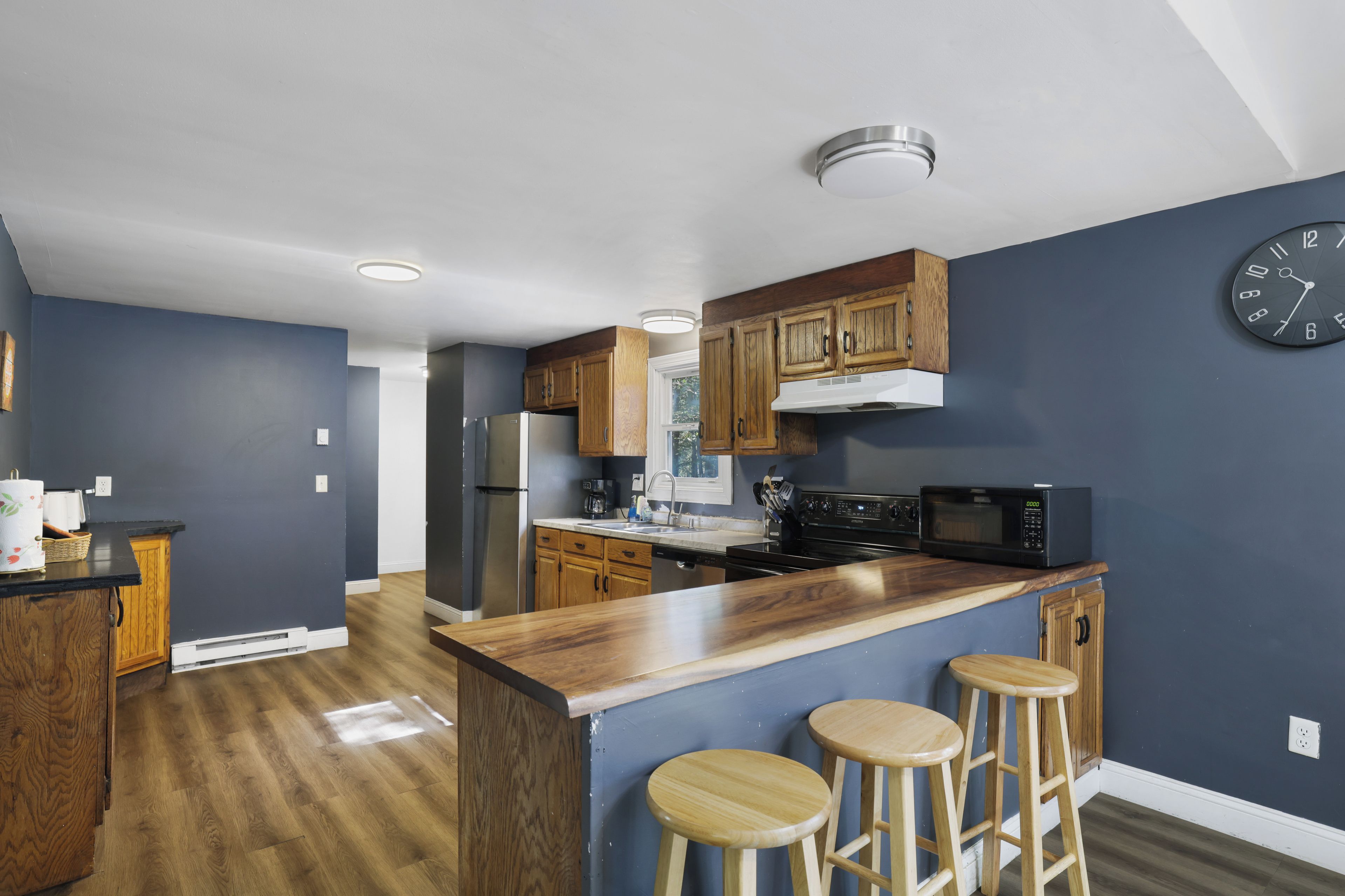 A modern kitchen with wooden cabinets, a stainless steel refrigerator, a microwave, and a bar area with stools.