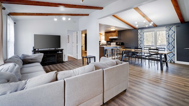 Una sala de estar moderna con un sofá seccional, un televisor de pantalla plana, vigas de madera y una zona de cocina con taburetes de bar.