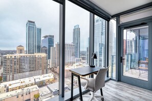 Enjoy a panoramic city view from the office space with a large window, minimalist desk, and glass door leading to a balcony.