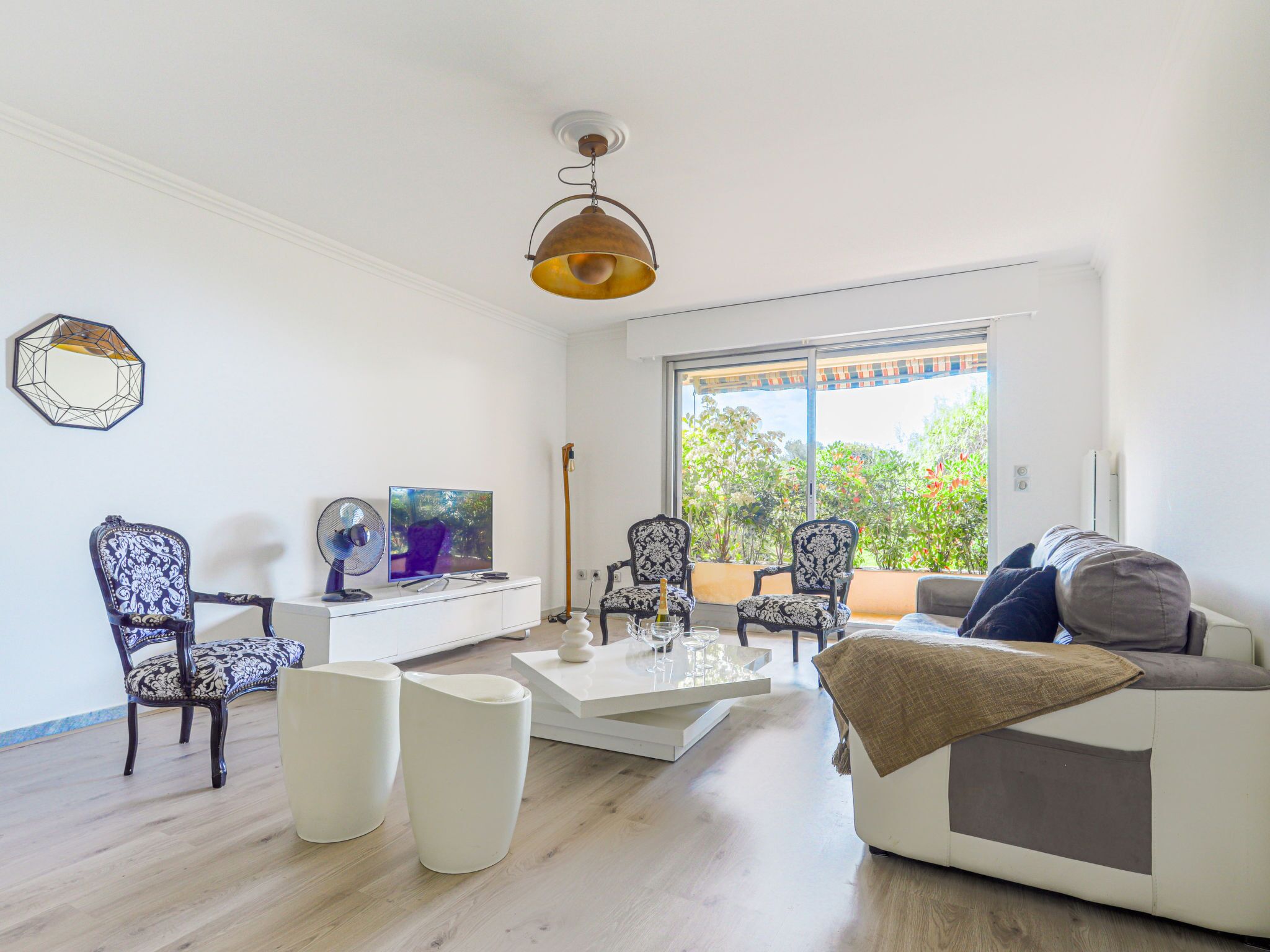 A modern living room with a white sofa, two patterned chairs, a coffee table, a TV, and a large window with a view of greenery.
