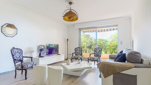 A modern living room with a white sofa, two patterned chairs, a coffee table, a TV, and a large window with a view of greenery. - Les Balcons d'Eze by Interhome (Eze)
