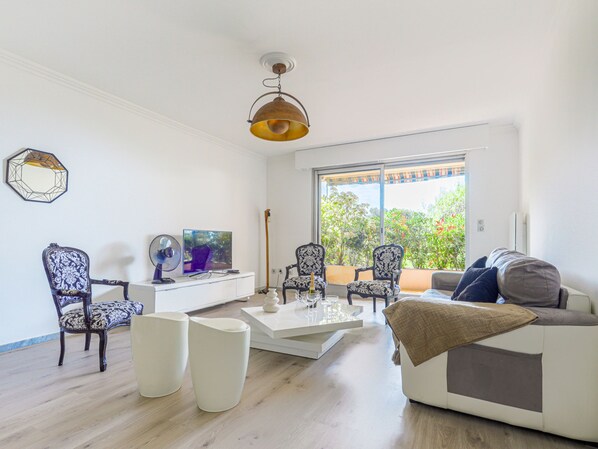 A modern living room with a white sofa, two patterned chairs, a coffee table, a TV, and a large window with a view of greenery.