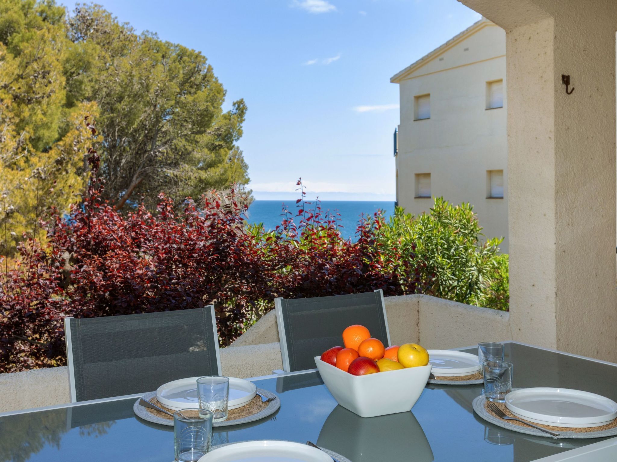An outdoor dining table with a bowl of fruit, plates, and glasses, overlooking a body of water and trees.