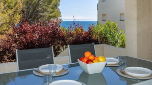 Une table à manger en plein air, avec un saladier de fruits, des assiettes et des verres, donnant sur une étendue d’eau et des arbres.