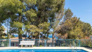 Une piscine d’un bleu éclatant, avec un banc en bois et une rambarde en métal, entourée d’arbres et offrant une vue sur la mer.