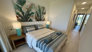 Une chambre à coucher avec un grand lit, deux tables de chevet avec des lampes, une œuvre d’art murale et une vue sur l’extérieur par une porte ouverte.