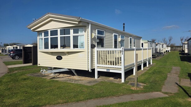 A mobile home with a front porch and a garage, situated in a grassy area with a clear sky. - Caravan Sleeps 6 With Game Console (Skegness)