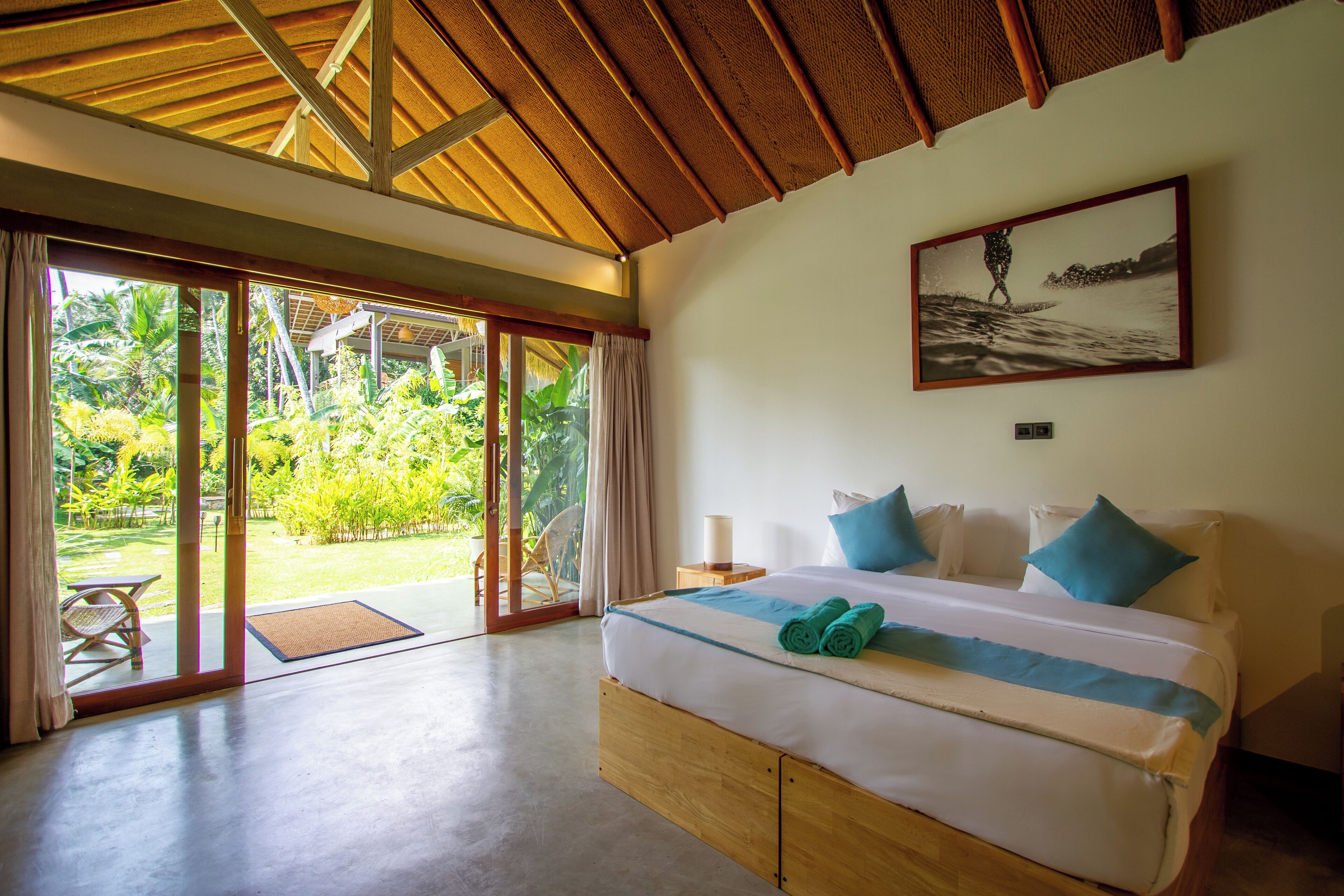 A spacious bedroom with a large bed, a view of a tropical garden through sliding glass doors, and a ceiling with exposed wooden beams.