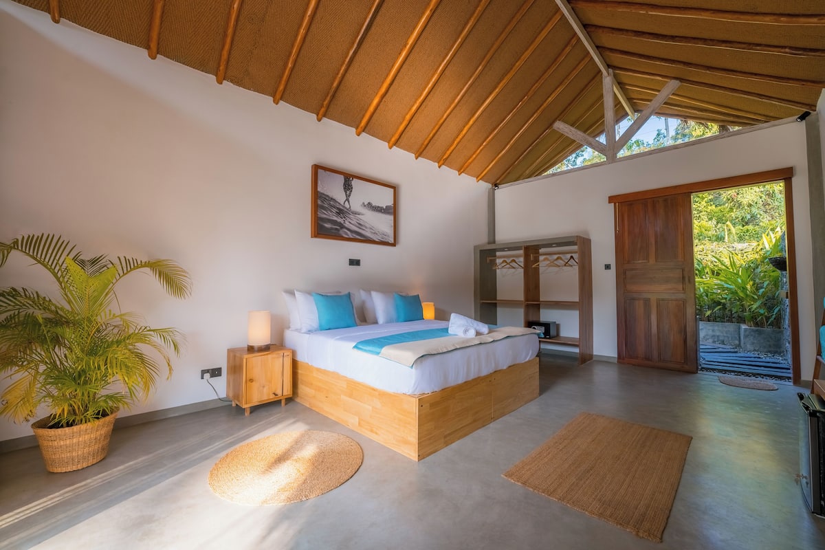 A bedroom with a large bed, a potted plant, a wooden cabinet, and a view of greenery outside.