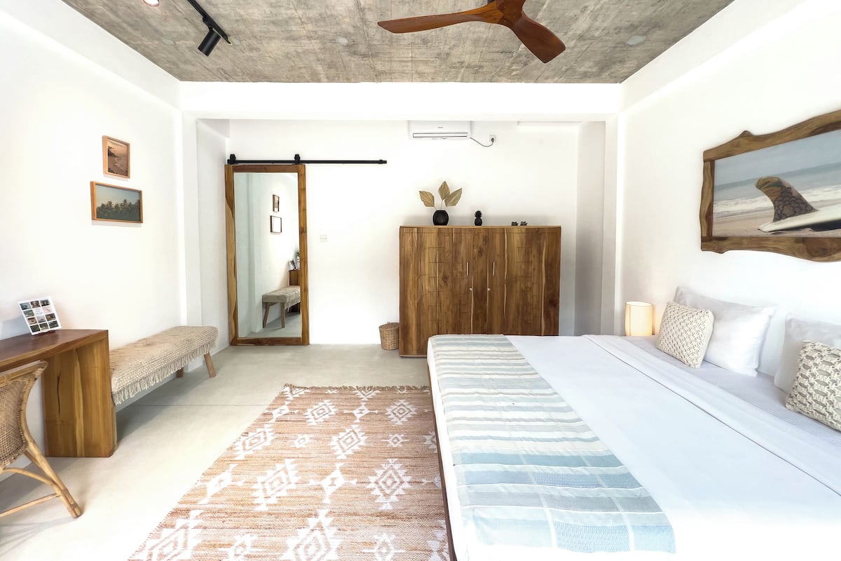 A bedroom with a large bed, a wooden wardrobe, a bench, a desk, and a ceiling fan.