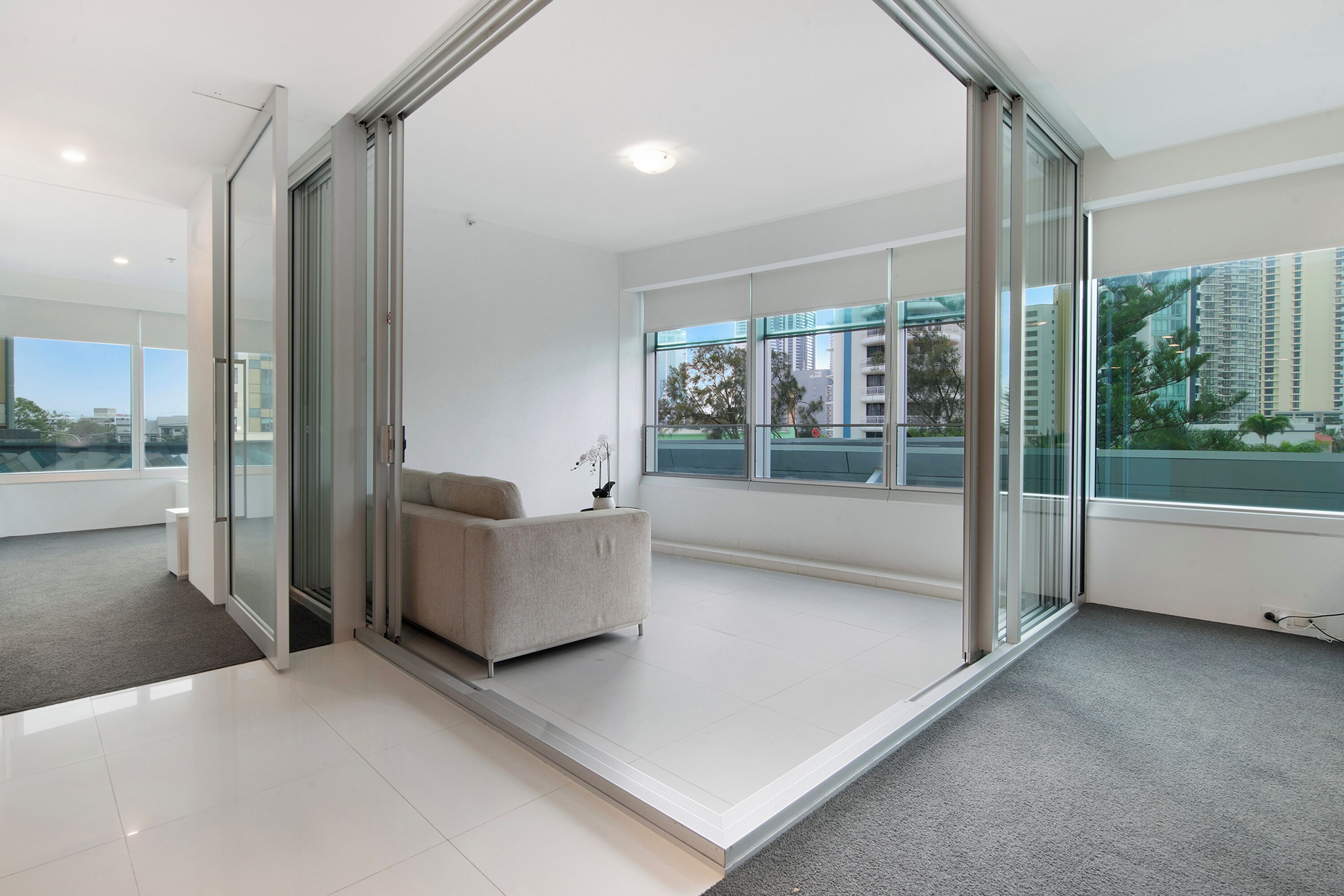 A modern hotel room with large glass doors leading to a balcony, a sofa, and a view of the cityscape.